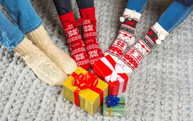 Three people wearing Christmas socks touching their feet to a pile of presents